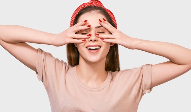 Foto fröhliche junge frau, die ihr gesicht mit den fingern versteckt, die ihr auge bedecken hübsche frau, die ihr gesicht von hand mit beigefarbenem t-shirt, rotem stirnband und roten nägeln mit geschlossenen augen bedeckt, die auf einer weißen wand posieren