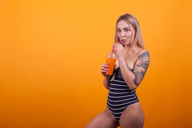 Fröhliche junge Frau, die köstlichen orangefarbenen Cocktail in ihrem sexy Badeanzug trinkt und die Kamera über gelbem Hintergrund betrachtet. Mädchen mit attraktivem Körper. Schöne Frau im Badeanzug.
