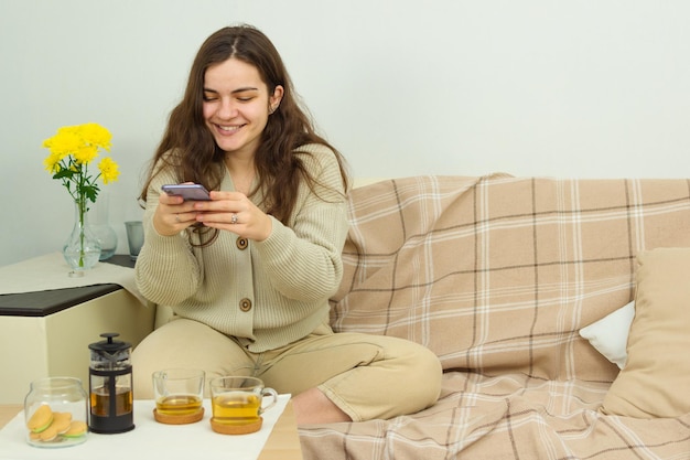 Fröhliche junge Frau, die lächelt und Fotos auf dem Handy von Tassen mit Tee auf dem Tisch macht