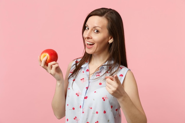 Fröhliche junge Frau, die mit dem Zeigefinger auf die Kamera zeigt und frische reife rote Apfelfrüchte isoliert auf rosa Pastellwandhintergrund hält. Lebendige Lebensweise der Menschen, entspannendes Urlaubskonzept. Mock-up-Kopienbereich.