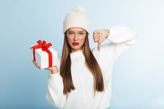 Fröhliche junge frau, die pullover und hut trägt, geschenkbox hält und daumen nach unten zeigt