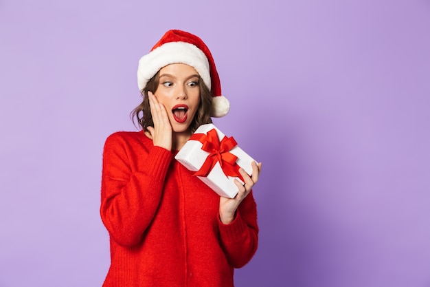 Fröhliche junge Frau, die roten Weihnachtshut trägt, der lokal über violetter Wand steht und Geschenkbox hält