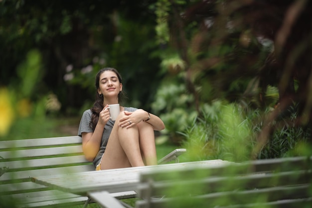 Fröhliche junge Frau, die sich entspannt und frische Luft atmet, während sie auf dem Balkon mit Blick auf die Natur sitzt und morgens Kaffee und Getränke trinkt