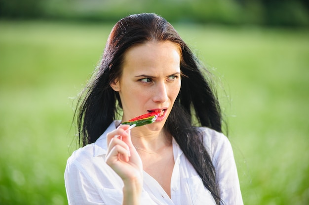 Fröhliche junge frau, die süßigkeiten in form von wassermelonenscheiben nahe ihrem gesicht hält. mädchen, das in der natur mit lutschern ruht.