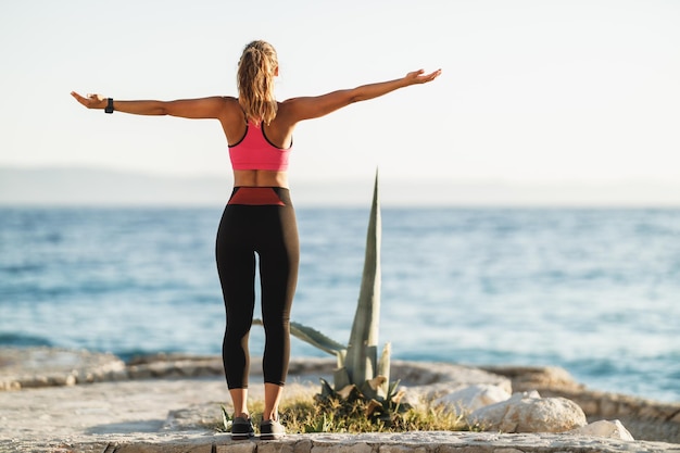 Fröhliche junge Frau, die tiefe frische Luft einatmet und die Arme beim Stretching-Training in der Nähe des Meeresstrandes hebt.