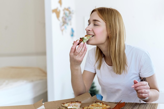 Fröhliche junge Frau, die zu Hause ein Stück heiße Pizza isst und eine köstliche Mahlzeit genießt