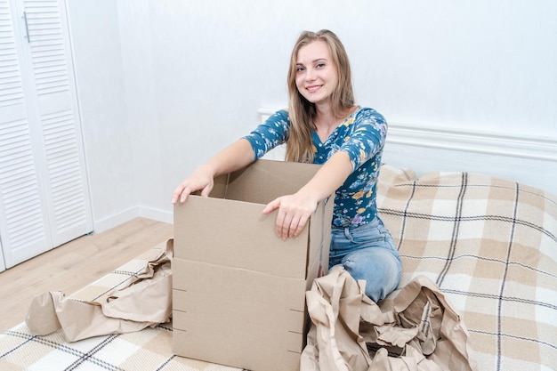 Fröhliche junge Frau, die zu Hause mit offenem Karton auf dem Bett sitzt Paket auspacken