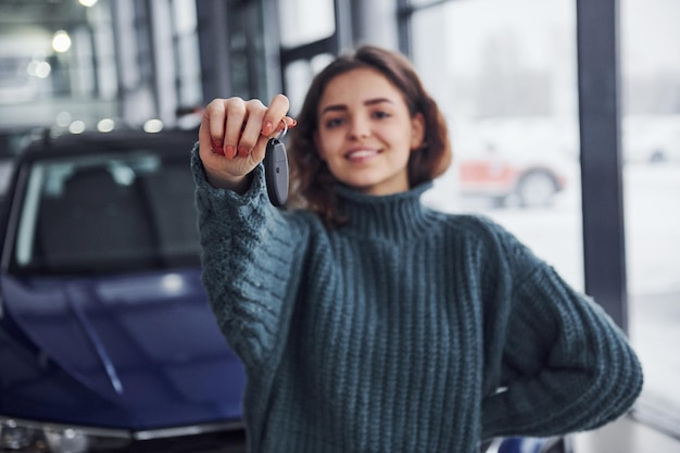Fröhliche junge Frau hält Schlüssel und steht drinnen vor modernem Neuwagen.