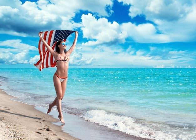 Fröhliche junge Frau im Bikini, die mit der Flagge am Strand läuft