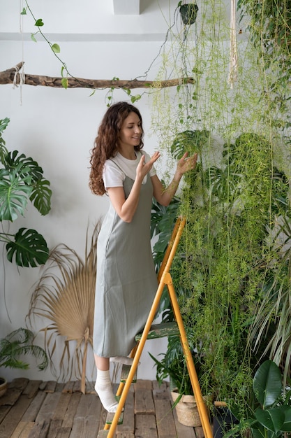 Fröhliche junge Frau im Kleid, die grüne Blätter im heimischen Gartengewächshaus-Dschungel berührt