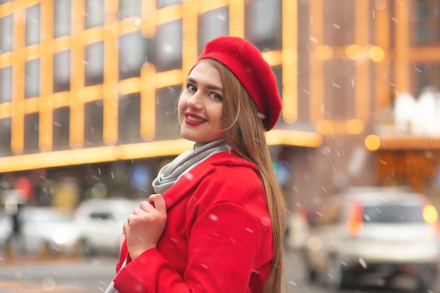 Fröhliche junge Frau im roten Mantel, die während des Schneefalls auf dem Straßenfest spazieren geht