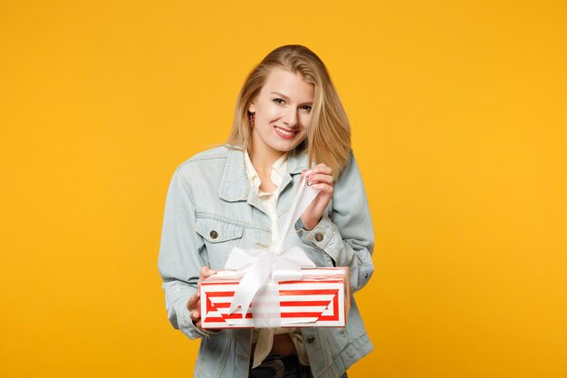 Fröhliche junge Frau in Denim-Freizeitkleidung hält rot gestreifte Geschenkbox mit Geschenkband isoliert auf gelb-orangefarbenem Hintergrund. St. Valentinstag Internationaler Frauentag Geburtstag, Urlaubskonzept.