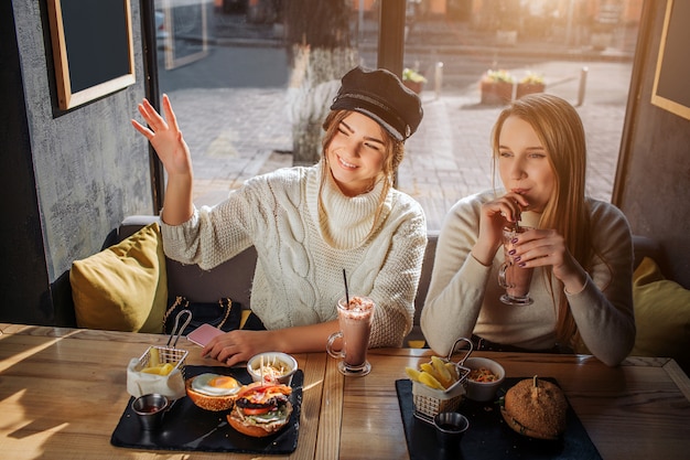 Fröhliche junge Frau in der Mütze sitzen mit ihrer Freundin am Tisch. Sie winkt mit der Hand und lächelt. Zweites Modell Getränkecocktail. Sie haben Essen am Tisch.