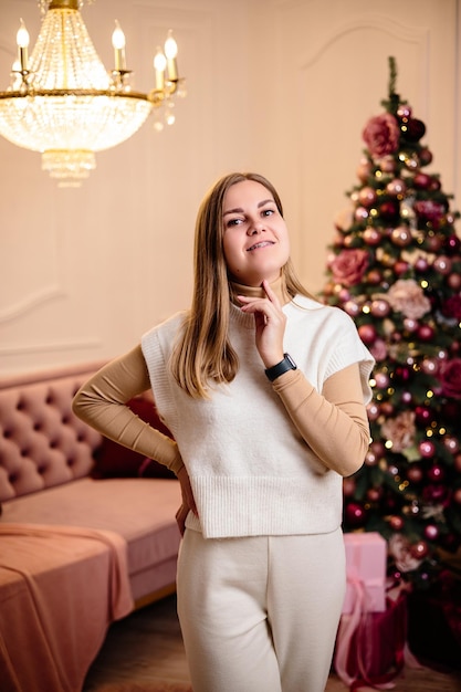 Fröhliche junge Frau in einem weißen Strickanzug auf dem Hintergrund festlicher Weihnachtsbäume Atmosphäre des neuen Jahres