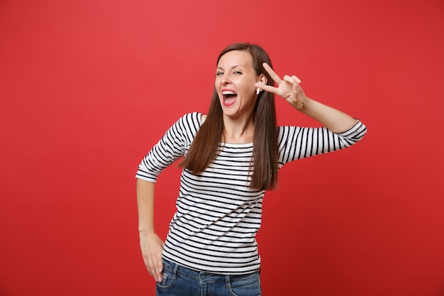 Fröhliche junge Frau in gestreifter Kleidung mit drahtlosen Kopfhörern mit Victory-Zeichen, Musik hören isoliert auf rotem Wandhintergrund. Menschen aufrichtige Emotionen, Lifestyle-Konzept. Kopieren Sie Platz.