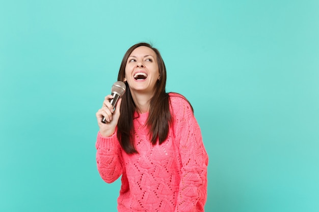 Fröhliche junge Frau in gestricktem rosa Pullover, die in der Hand nach oben schaut, ein Lied im Mikrofon singt, das auf blautürkisem Wandhintergrund isoliert ist, Studioporträt. Menschen Lifestyle-Konzept. Kopieren Sie Platz.