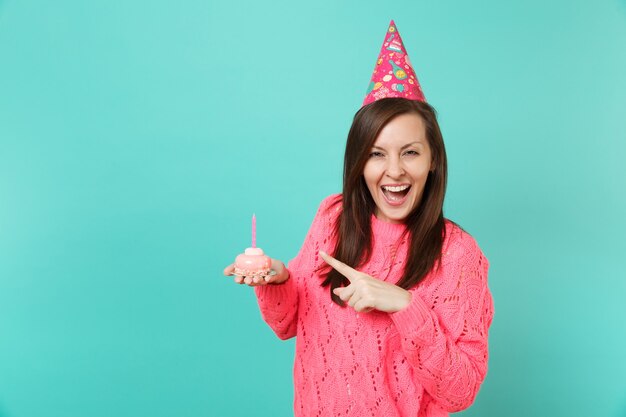 Fröhliche junge Frau in gestricktem rosa Pullover, Geburtstagshut zeigt Zeigefinger auf Kuchen mit Kerze in der Hand isoliert auf blauem Hintergrund Studioporträt. Menschen Lifestyle-Konzept. Kopieren Sie Platz.