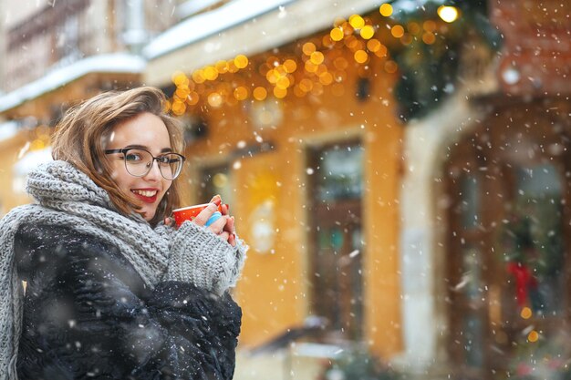 Fröhliche junge Frau in Gläsern mit einer Tasse Kaffee, die bei Schneewetter in der Nähe von dekorierten Vitrinen spaziert. Freiraum