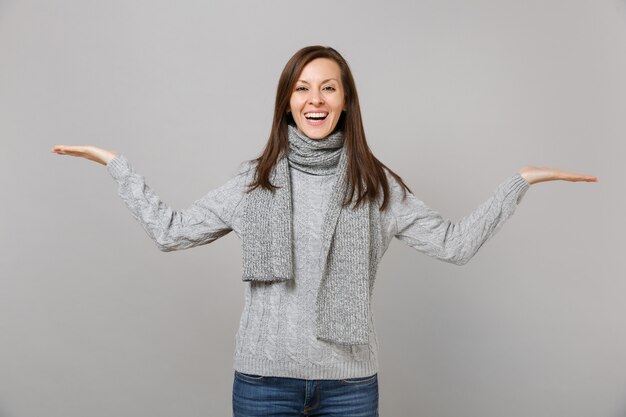 Fröhliche junge Frau in grauem Pullover, Schalausbreitung, Hände beiseite zeigend einzeln auf grauem Wandhintergrund. Gesunder Modelebensstil, aufrichtige Emotionen der Menschen, Konzept der kalten Jahreszeit. Kopieren Sie Platz.