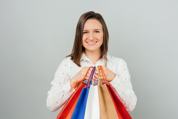 Fröhliche junge Frau in lässiger Kleidung mit bunten Einkaufstaschen auf grauem Hintergrund