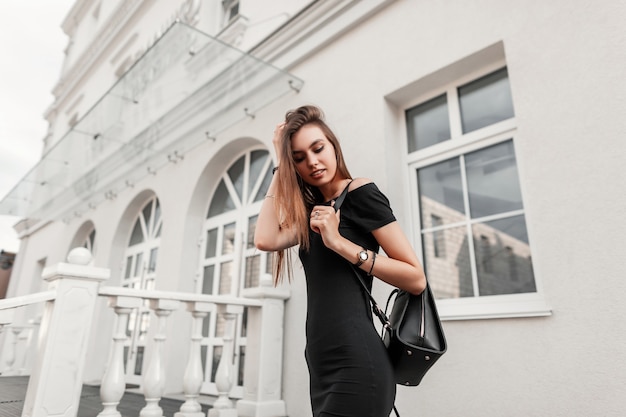 Fröhliche junge Frau in schwarzer modischer Kleidung glättet Haare. Attraktives freudiges Mädchenmodell in einem schwarzen Kleid mit einem Rucksack wirft nahe einem weißen Gebäude im Freien auf. Lässiger Herbstlook.