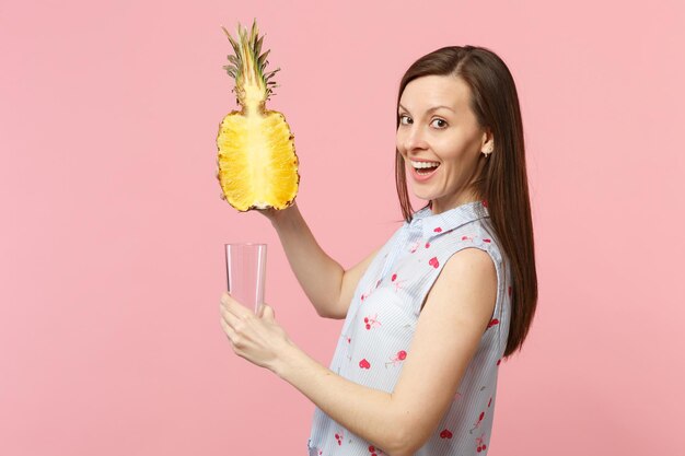 Fröhliche junge Frau in Sommerkleidung, die die Hälfte der frischen reifen Ananas-Fruchtglasschale isoliert auf rosa Pastellwandhintergrund hält. Lebendige Lebensweise der Menschen, entspannendes Urlaubskonzept. Mock-up-Kopienbereich.