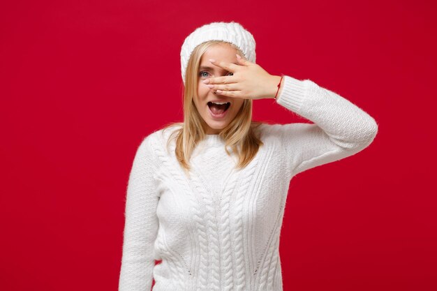 Foto fröhliche junge frau in weißer strickmütze isoliert auf rotem hintergrund studioportrait. gesunde mode lifestyle menschen emotionen konzept der kalten jahreszeit. mock-up-kopienbereich. bedecken sie die augen mit der hand, die sich versteckt.