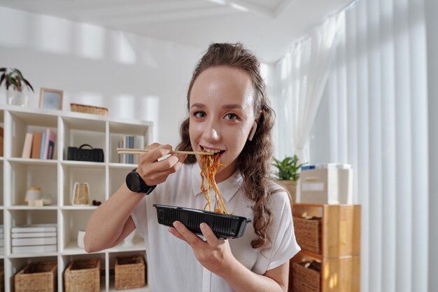 Fröhliche junge Frau isst Nudeln zum Mittagessen in ihrem Homeoffice