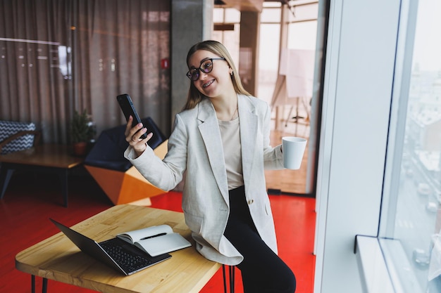 Fröhliche junge Frau lacht, während sie lustige Benachrichtigungen auf dem Smartphone liest, das vor dem Laptop im Café sitzt Glückliche Frau, die Geld über die Internet-Banking-App auf dem Handy sendet