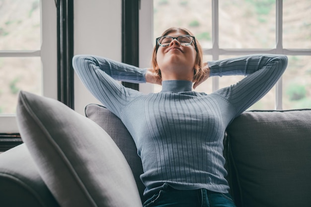 Fröhliche junge Frau mit Brille, die sich auf dem Sofa mit den Händen hinter dem Kopf im Wohnzimmer ihres Hauses entspannt Sorglose und zufriedene Dame beim Entspannen auf der Couch im modernen apartmentxA