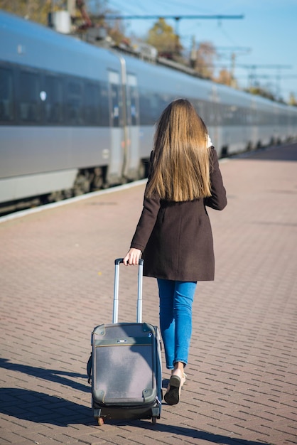 Fröhliche junge Frau mit Brille im Mantel mit großer Reisetasche am Bahnhof auf Zughintergrund