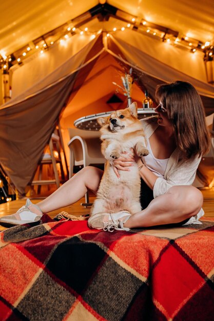 Fröhliche junge Frau mit ihrem walisischen Corgi Pembroke-Hund, der sich am Sommerabend in der Nähe des gemütlichen Lagerfeuers beim Glamping entspannt Luxus-Campingzelt für Erholung im Freien und Erholung Lifestyle-Konzept