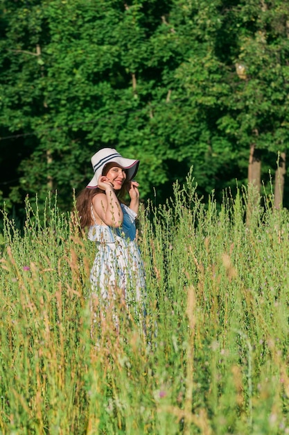 Fröhliche junge Frau mit langen Haaren in Hut und Kleid, die an einem sonnigen Tag durch den Sommerwald geht Sommerfreude-Konzept