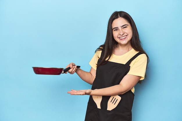 Fröhliche junge Frau mit Pfanne auf einem leuchtend blauen Hintergrund