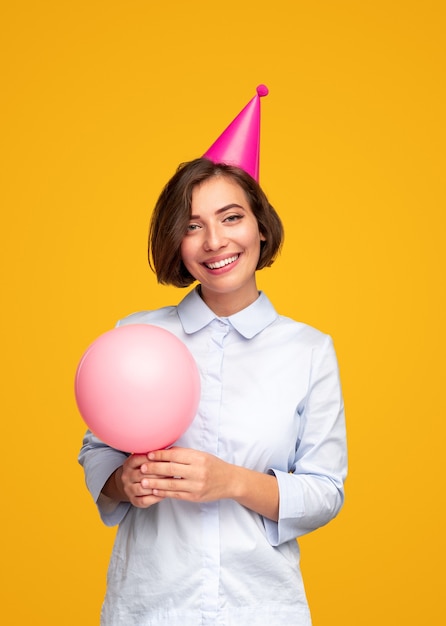 Fröhliche junge Frau mit rosa Ballon lächelnd und Kamera betrachtend, während Geburtstag gegen gelben Hintergrund feiern