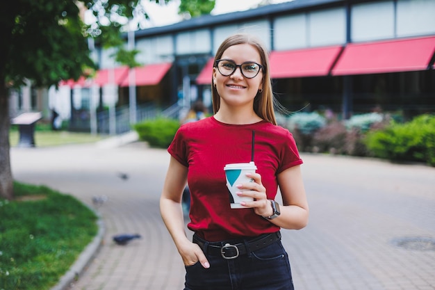 Fröhliche junge Frau mit Rucksack, die lächelt und wegschaut, während sie auf der Straße der Stadt spazieren geht und frischen Kaffee zum Mitnehmen genießt
