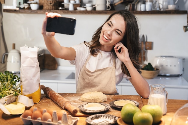 Fröhliche junge Frau mit Schürze bereitet Teig für einen Apfelkuchen in der Küche zu Hause vor und macht ein Selfie