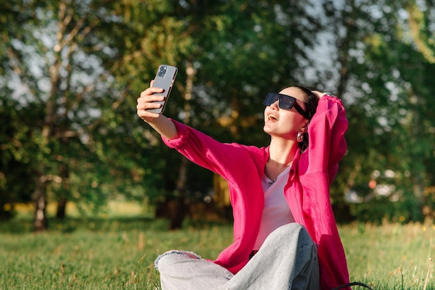 Fröhliche junge Frau mit Sonnenbrille und Handy, die an sonnigen Tagen auf Gras im Park sitzt Frau fotografiert sich selbst