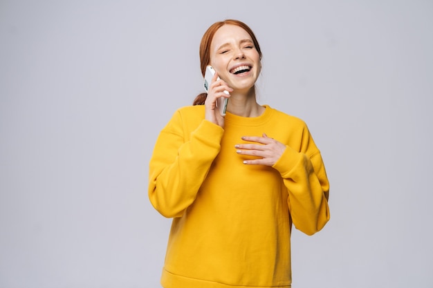 Fröhliche junge frau mit stylischem gelben pullover, die mit dem handy auf isoliertem weißem hinterg...