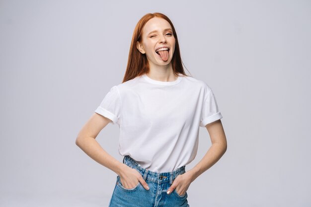 Fröhliche junge Frau mit T-Shirt und Jeans, die Zunge auf isoliertem weißem Hintergrund zeigt