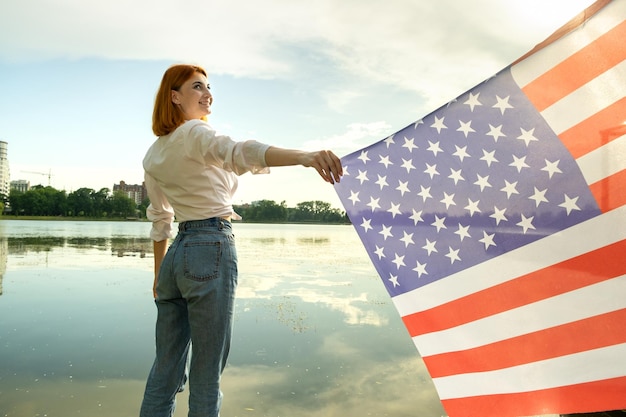 Fröhliche junge Frau mit US-Nationalflagge auf ihren Schultern mit hohen Stadtgebäuden im Hintergrund, die den Unabhängigkeitstag der Vereinigten Staaten feiern