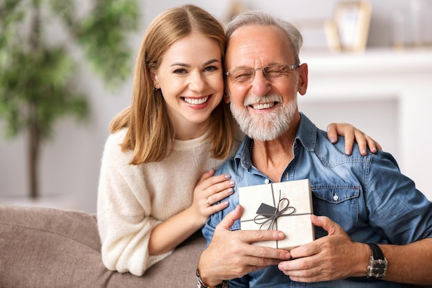 Fröhliche junge Frau umarmt älteren Mann, während sie auf der Couch sitzt und Geschenkbox während der Feiertagsfeier zu Hause öffnet