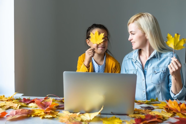 Fröhliche junge Frau und kleines Mädchen, die online arbeiten, Webinar ansehen, Podcasts auf dem Laptop ansehen und sich zu Hause um Herbstblätter herum unterhalten