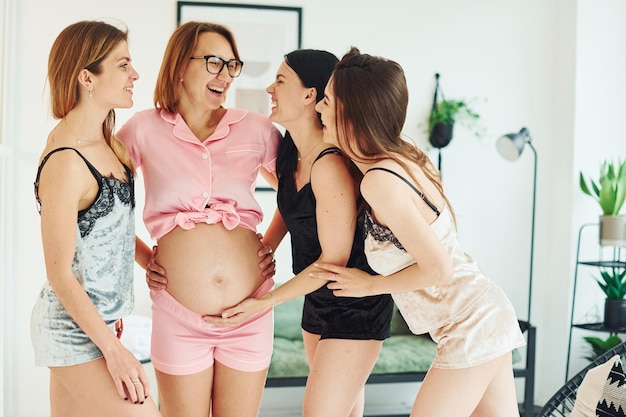 Fröhliche junge Frauen mit ihrer schwangeren Freundin im Schlafanzug stehen und feiern tagsüber gemeinsam drinnen