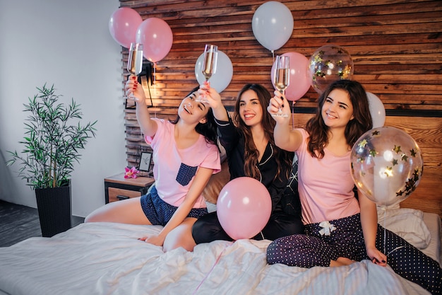 Fröhliche junge Frauen sitzen auf dem Bett im festlichen Raum. Sie halten Gläser mit Champagner in die Luft und schauen sie an. Mädchen lächelnd. Sie haben Pyjamas Party.