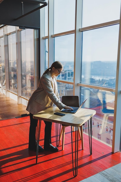 Fröhliche junge Freiberuflerin in Freizeitkleidung, die mit einem Netbook an einem Holztisch steht und Notizen schreibt, während sie an einem Projekt arbeitet Moderne junge Geschäftsfrau im Büro