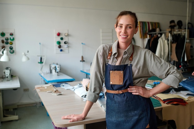 Fröhliche junge Handwerkerin in Schürze, die am Arbeitsplatz im Atelier steht