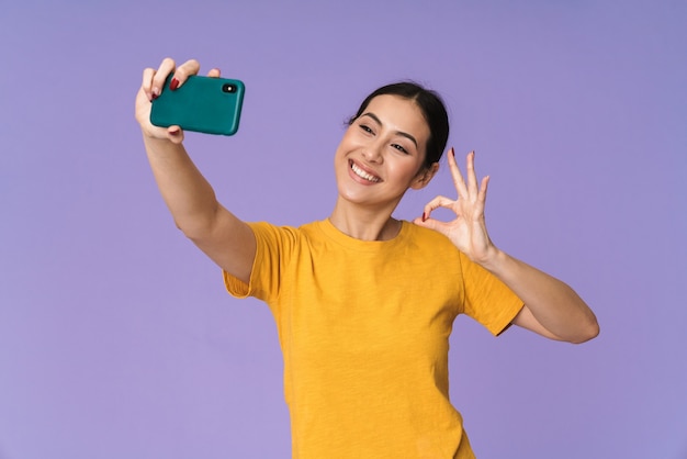 Fröhliche junge hübsche Sportlerin, die isoliert über der violetten Wand steht, ein Selfie macht und eine ok Geste zeigt