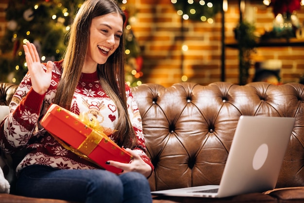 Fröhliche junge kaukasische Dame sitzt auf einem Couch-Chat in der Videokonferenz-App auf der Laptop-Show-Geschenkbox