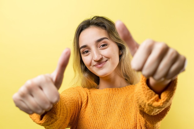 Fröhliche junge kaukasische Frau in einem orangefarbenen Pullover, die ein Daumenzeichen macht und lächelt Gute Arbeit und Respekt